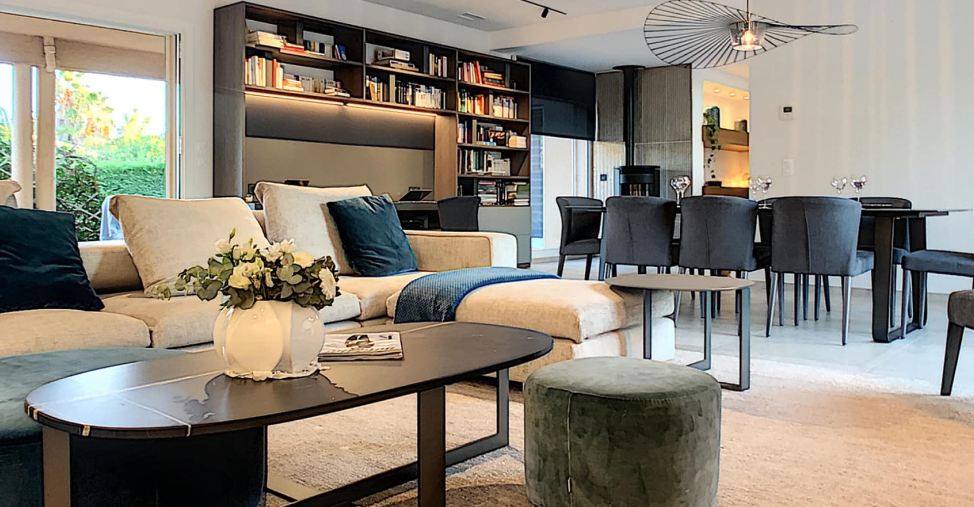 Salon moderne et lumineux avec canapé beige, bibliothèque encastrée éclairée et salle à manger grise adjacente, ambiance cosy.